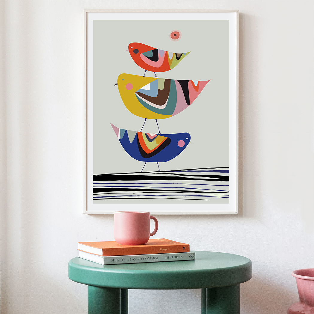Colorful abstract bird artwork on a wall above a green table with books and a pink mug.
