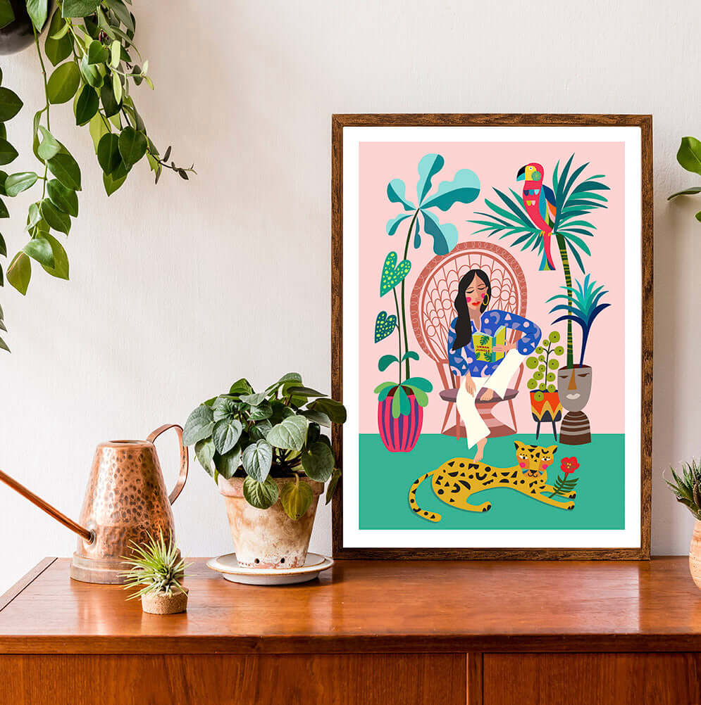 The Print featuring a Woman is reading a book on a chair and leopard chilling on the floor. this print on a table surrounded by plants.