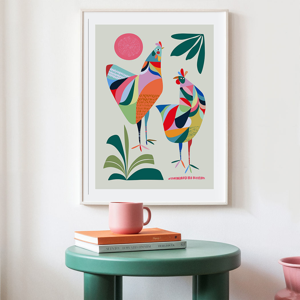 Colorful abstract art print of two birds on a wall above a green table with books and a mug.