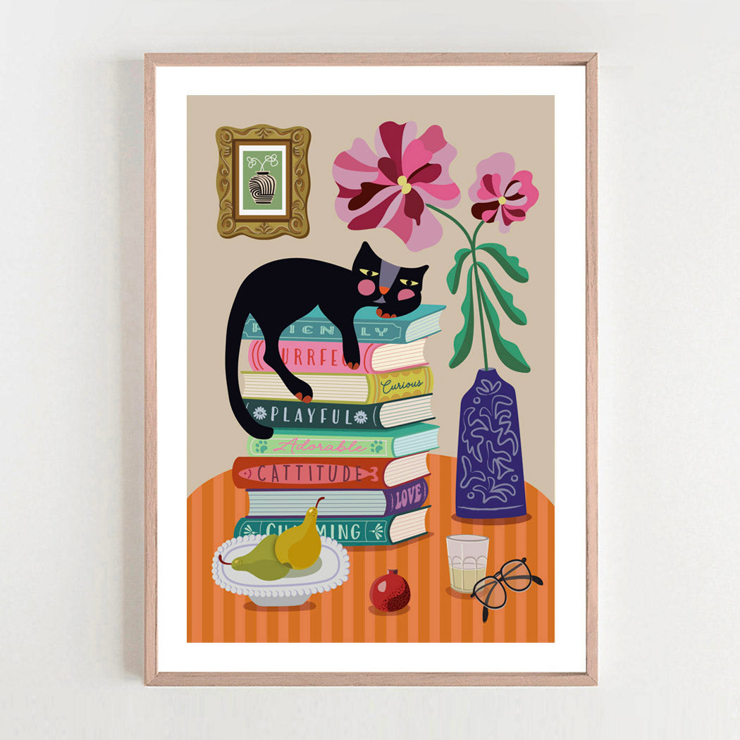 Framed artwork with a black cat on books, flowers, and a vase on a table.