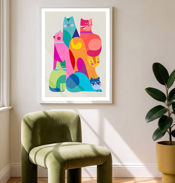 A framed wall art print featuring a colorful stack of stylized cats in bright pink, teal, orange, yellow, and green tones, hung on a beige wall above an olive green velvet chair with a potted plant nearby.