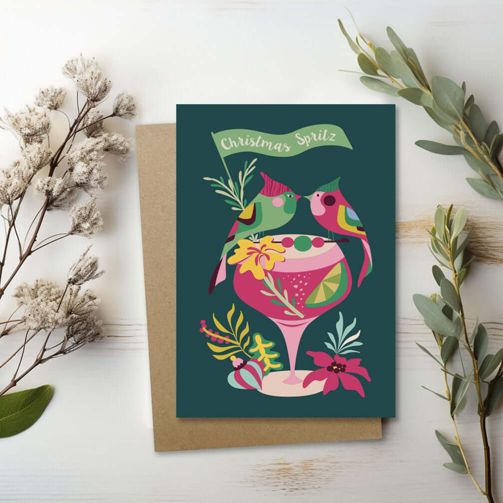 A greeting card featuring 2 colorful birds standing on a cocktail glass and surrounded by vibrant flowers.