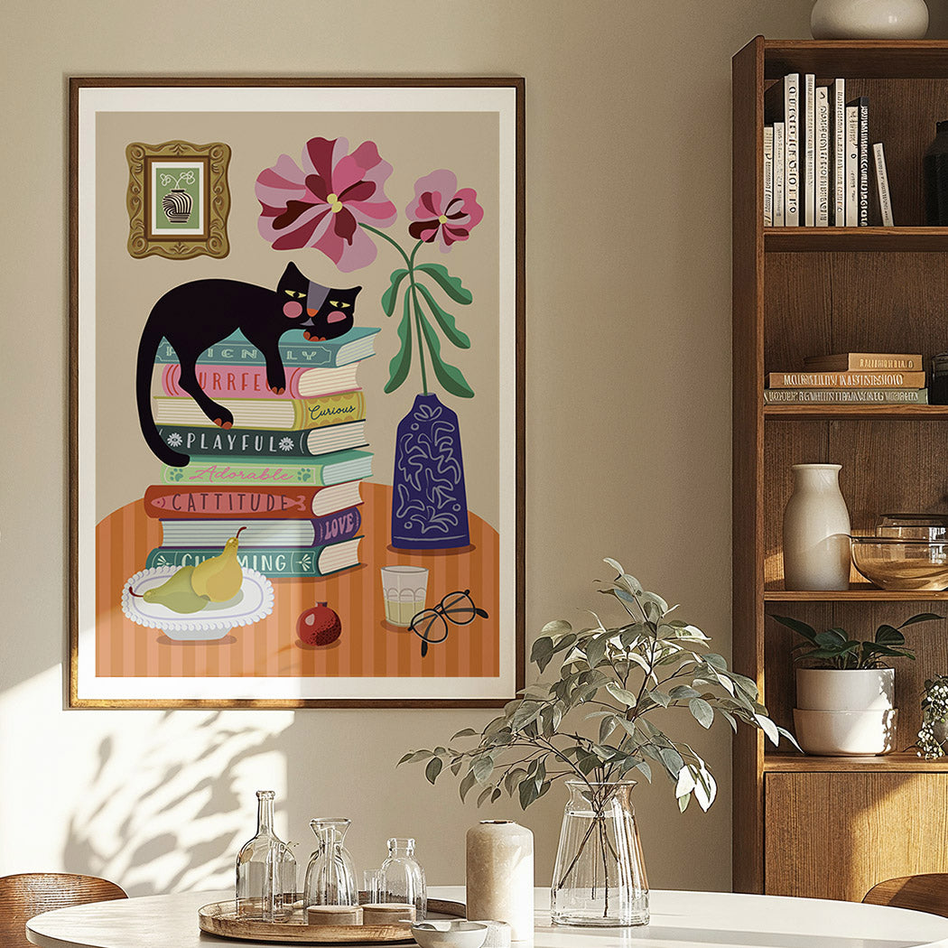 Framed artwork of a cat on books with flowers and a vase, displayed in a room with a bookshelf and table.