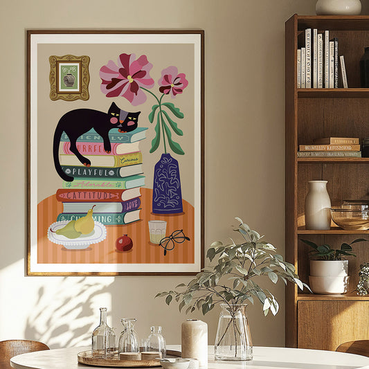 Framed artwork of a cat on books with flowers and a vase, displayed in a room with a bookshelf and table.