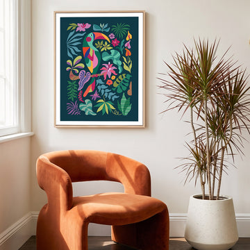 A framed tropical art print featuring a colorful toucan and lush foliage, styled on a light beige wall above a rust-orange velvet armchair and next to a potted plant.